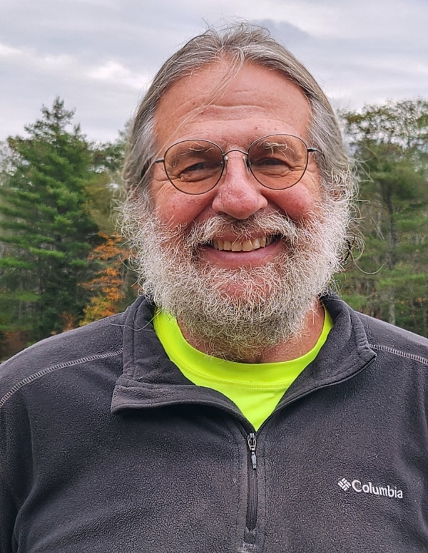 A man with long salt and pepper hair pulled back and a full beard smiles at the camera. He wears wire-frame glasses and has light skin. He is wearing a bright neon green t-shirt beneath a gray fleece jacket. Green pines and autumn foliage are in the background.