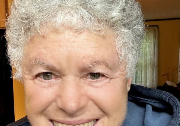 A woman with short, curly silver hair smiles at the camera. She has light-toned skin and is wearing a cloudy blue hoodie. Shelves and a curtained window are in the background.