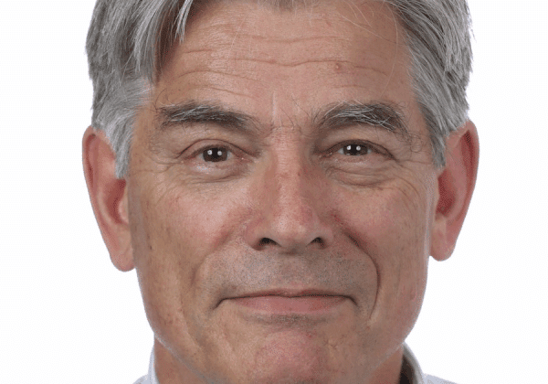A man with salt and pepper hair smiles past the camera. He has light skin and is wearing a pale blue button-down shirt.