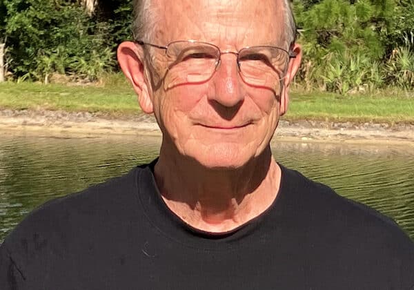 A man with short-cropped silver hair and light skin smiles past the camera. He is wearing wire-frame glasses and a black crew-neck shirt. Green palms and a small body of water are in the background.