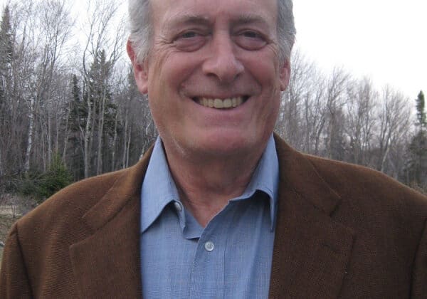 Aman with silver hair at the temples and light skin smiles at the camera. He is wearing a blue button-down shirt and a brown wool blazer. Bare trees and a gray sky are in the background.