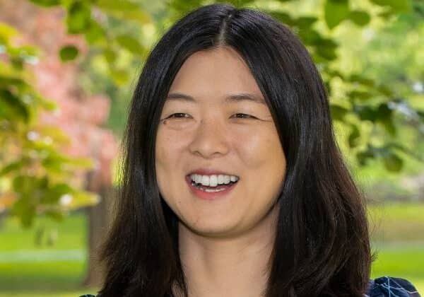 A woman with shoulder length black hair smiles at the camera. She has medium-toned skin and is wearing a black blouse with white swirl patterns. Green trees and grass are in the background.