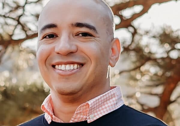 A man with light-medium toned skin and dark eyes smiles at the camera. He is wearing a light pink collared shirt under a navy blue sweater. Autumn trees are in the background.