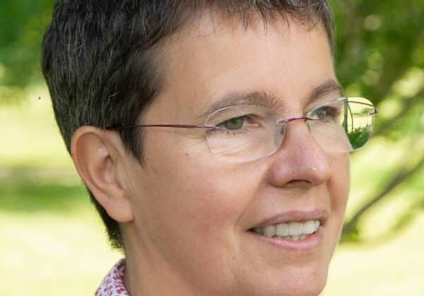 A woman with short-cropped brown hair smiles into the distance. She has light-toned skin and is wearing frameless glasses. Her collared shirt has a red pattern on white. Green trees and grass are in the background.