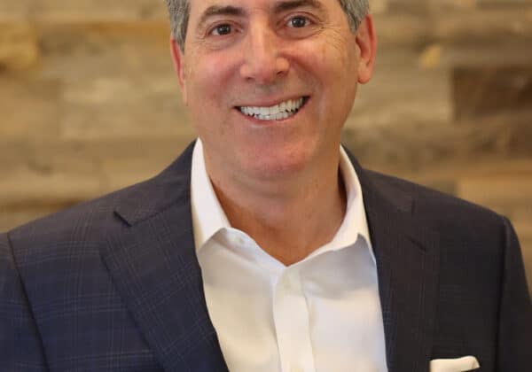 A man in a dark suit and white dress shirt smiles at the camera. He has short salt and pepper hair and light-toned skin. A wall of wooden boards is behind him