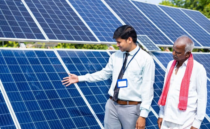 Two men stand in front of a set of solar panels. One is wearing a tie and dress shirt, he is explaining. The other man, listening, is wearing all white and an orange scarf.