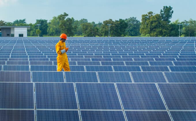 An inspector in an orange jumpsuit examines a field of solar panels.