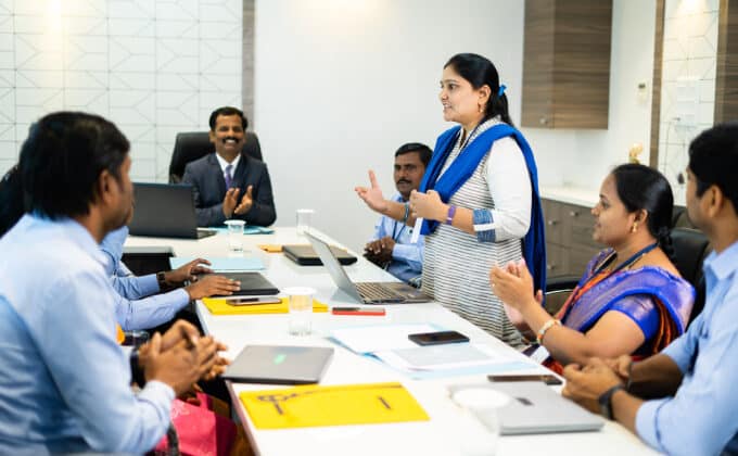 A group of decisionmakers sit around a table. One woman is standing and explaining to the others who listen intently.