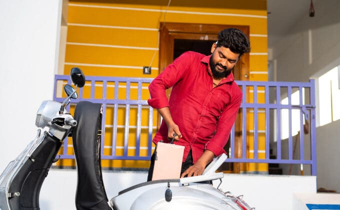 A man with black hair and a full beard is pulling the battery out of his electric scooter. He has medium-toned skin and is wearing a red dress shirt.