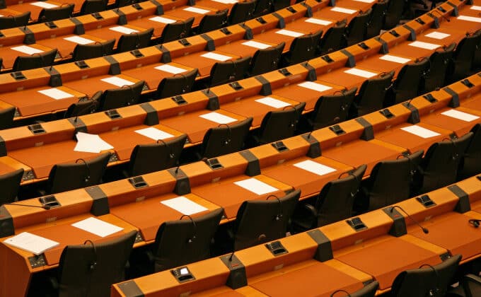 The empty seats and desks of a parliamentary room. Each desk has a sheet of paper and microphone.