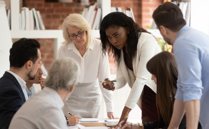 A group of decisionmakers around a table. A woman with dark skin leans over the table explaining to the others.