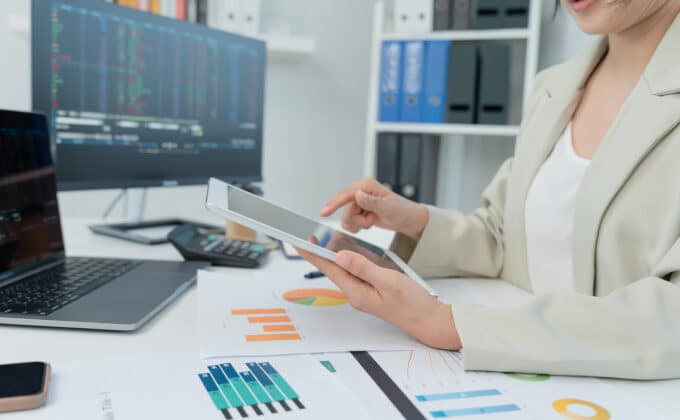 A woman with medium-toned skin is making calculations on a tablet. She is surrounded by analysis reports and her computer monitor shows stock investments.