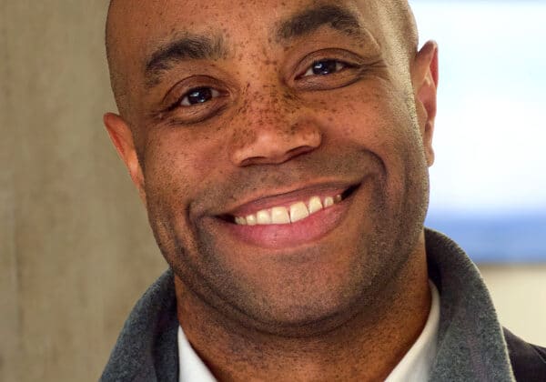 A man with a broad smile and medium-dark skin looks at the camera. He is wearing a white collared shirt and a grey, fleece zip-up under a navy blue blazer. Beige marble and a window are in the background.