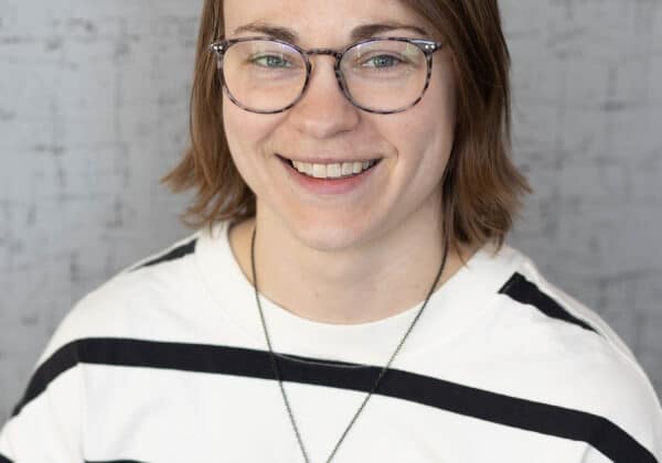 A woman with shoulder length ash blonde hair smiles at the camera. She has light skin and thin-framed glasses. She is wearing a white shirt with thick black horizontal stripes. The background is a grey wall.