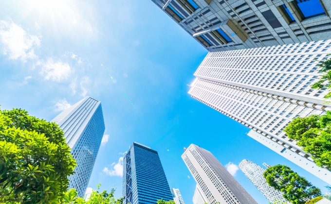 Modern office buildings climb toward a blue sky. Green trees edge the sides of the photo.