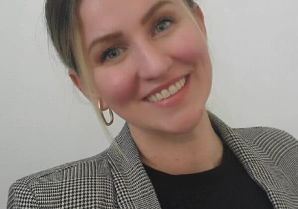 A woman with ash-blonde hair, pulled back, smiles at the camera. Her head is tilted to the left and she has light-toned skin. She is wearing a black shirt under a black and while checked blazer. The background is gray.