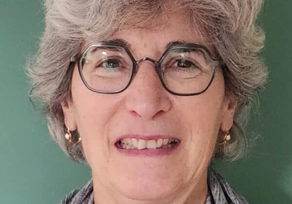 A woman with wavy salt and pepper hair smiles at the camera. She is wearing octagonal wire-framed glasses and a heather grey sweater with a cowl neck. The background is a sage green wall.