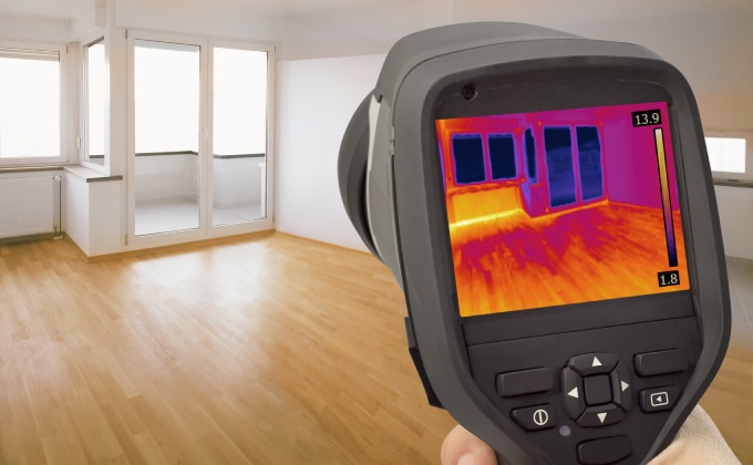 An energy efficiency meter with a digital reading of heat loss is in the foreground. An empty apartment is being scanned.