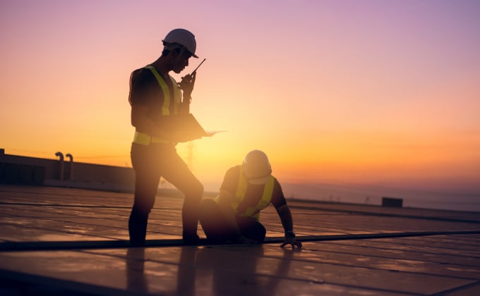 Two engineers stand working on a roof covered in solar panels. They are in silhouette against a colorful sunset.