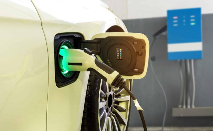 Close up of a white electric vehicle sedan. It is plugged in and the charger is glowing green.||