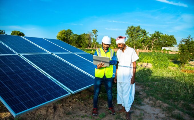 A young male technician worker wearing green vests and helmet checks the maintenance of the solar panels and talking about installation of new solar panel with farmer