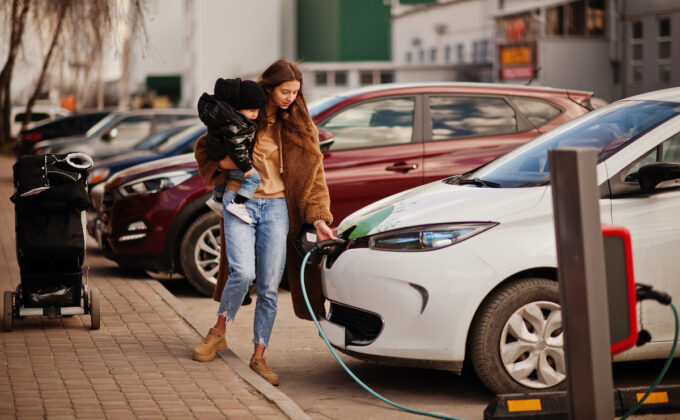 young woman with child on her hip plugs in electric vehicle along city street