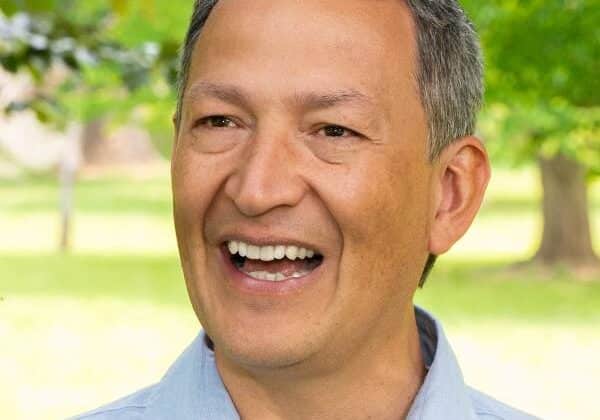 A medium skin tone man smiles at the camera. He has salt and pepper, short-cropped hair. He wears a pale blue collared shirt. Green trees and grass are in the background.