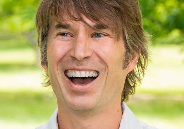 A man with light-toned skin laughs. He has long ash blonde hair swept across his forehead. He wears a white button-down shirt. Green trees and grass are in the background.