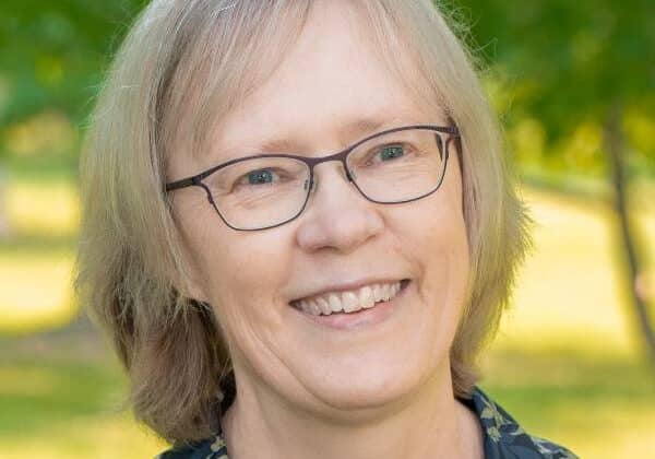 A woman with light skin and flax blonde hair cut just above her shoulders. She wears wire-framed glasses and a floral print shirt with collar. Green trees and grass are in the background.