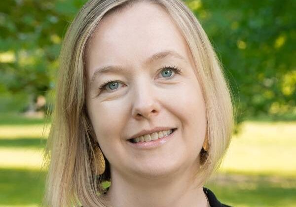 A woman with blue eyes and a blonde bob haircut, smiles at the camera. Her head is tilted to the left. She wears a black shirt and a silver wire necklace. Green trees and grass are in the background.