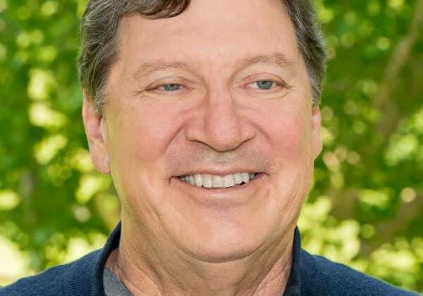 A man with wavy brown hair, flexed with gray, smiles at the camera. He has b;ue eyes and is wearing a navy blue zip-up sweater over a grey t-shirt. Green trees are in the background.