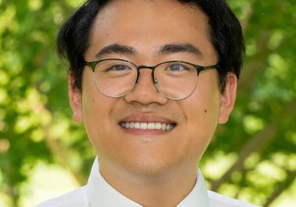 A man with medium-toned skin and short, black hair smiles past the camera. He is wearing a white button-down shirt, a dark tie, and wire-framed glasses. Green trees are in the background.