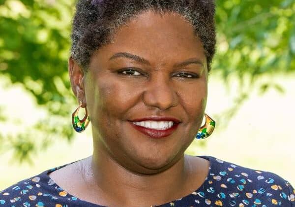 A woman with short-cropped curly hair and dark toned skin smiles at the camera. She wears a dotted shirt and beaded earrings. Green trees are in the background.