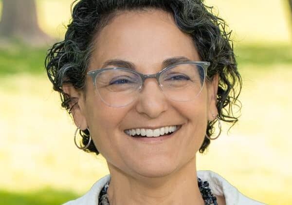 A woman with dark curly hair, cut short around her hears smiles past the camera. She has light-medium skin and wears wire-framed glasses. She is wearing a white jacket over a floral blouse. Green grass is in the background.