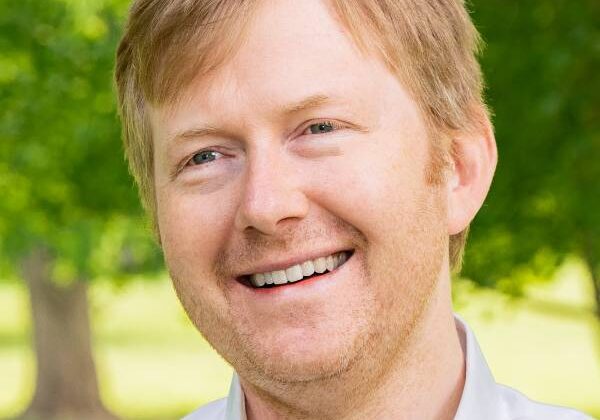 A man with short red hair and a beard smiles past the camera. He is wearing a checked collared shirt. Green trees and grass are in the background.