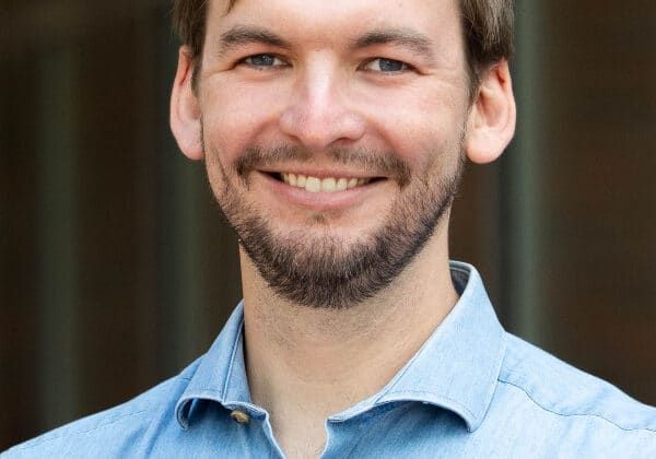 A man with brown hair swept to the right, a beard, and light skin smiles at the camera. He is wearing a light blue denim button-down. The background is blurred.