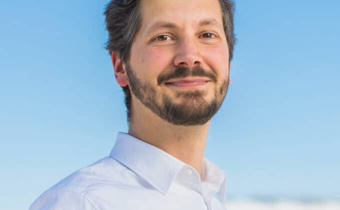 A man with dark short hair and a full beard smiles at the camera. He is wearing a white button down and has light skin. A cloudless blue sky fills the background.