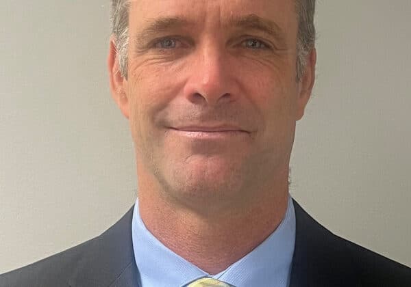 A man with short ash blonde hair smiles directly at the camera. He has light skin and is wearing a blue button down, yellow striped tie, and a dark blazer. The background is gray.