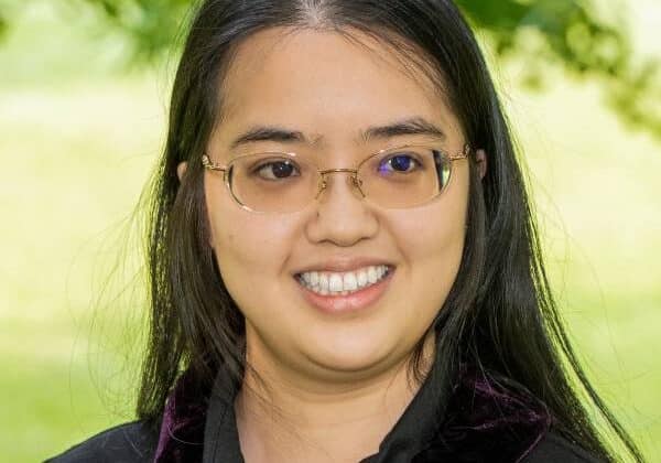 A woman with long, black hair smiles past the camera. She has medium-toned skin and is wearing a black collared shirt. She has frameless glasses. Green trees and grass are in the background.