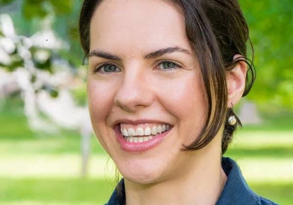 A woman with deep brown hair pulled back smiles broadly at the camera. She has light skin and is wearing a dark blue collared shirt. Green trees and grass are in the background.