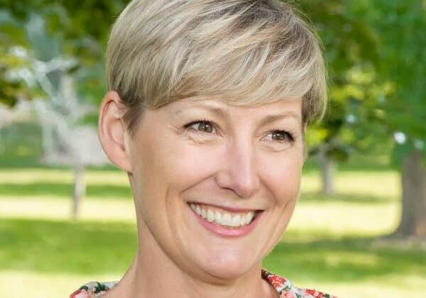 A woman with blonde hair cut short and bangs swept across her forehead smiles past the camera. She has brown eyes and light skin. She is wearing a floral, collared shirt. Green tress and grass are in the background.