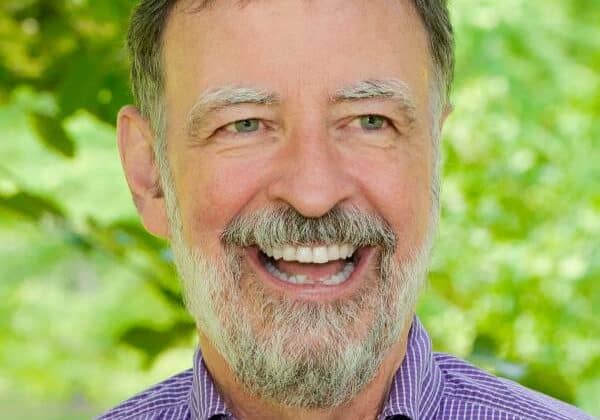 A man with short cropped dark hair and a full grey beard smiles broadly at the camera. He is wearing a purple checked button-down shirt. Green trees are in the background.