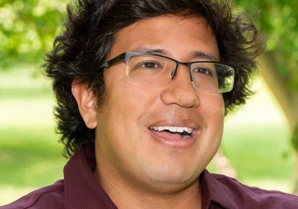 A man with wavy dark brown hair and medium-toned skin smiles past the camera. He is wearing frameless glasses and a burgundy collared shirt. Green trees and grass are in the background.