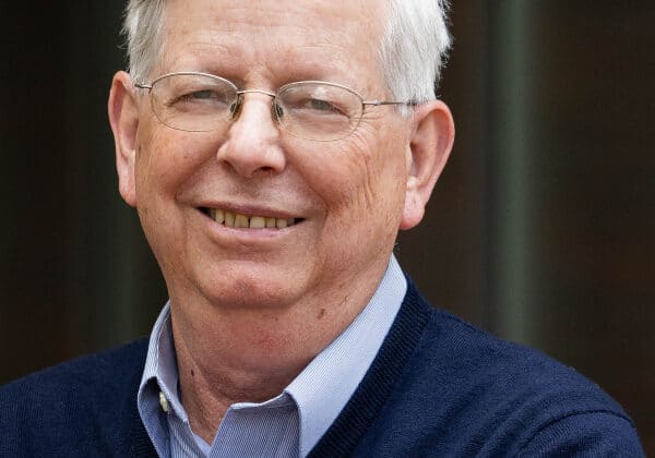 A man with silver hair swept to the right and light-toned skin smiles at the camera. He is wearing wire-framed glasses, a light blue collared shirt and a navy blue v-neck sweater. The background is blurred.