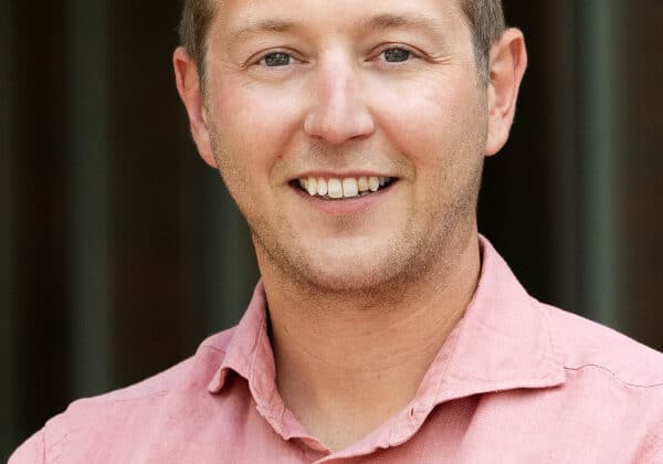 A man with wavy blonde hair and a slight beard smiles at the camera. He has light-toned skin and is wearing a salmon-colored button-down shirt. The background is blurred.