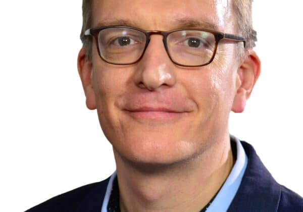 A man with short cropped blonde hair and light skin smiles at the camera. He is wearing dark framed glasses, a blue collared shirt, and a navy blazer. The background is white.