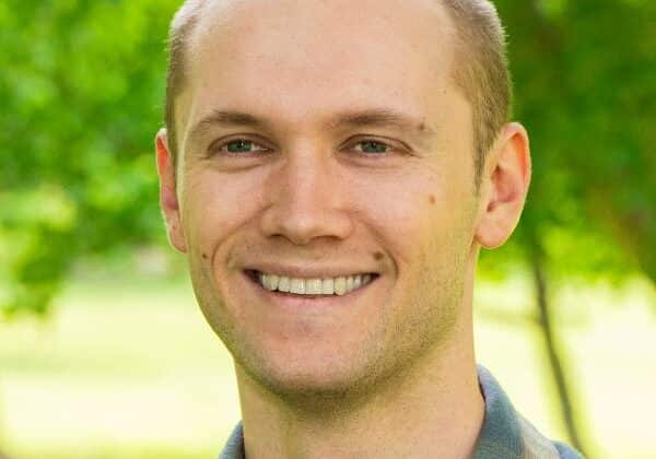 A man with buzz cut blonde hair and light skin smiles at the camera. He is wearing a blue and tan plaid button-down shirt. Green trees and grass are in the background.