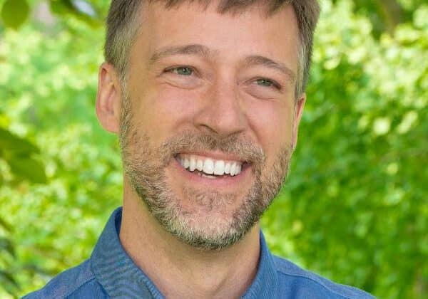 a man with short ash-blonde hair and a full beard smiles at the camera. He has light-skin and is wearing a blue collared-shirt. Green trees are in the background.