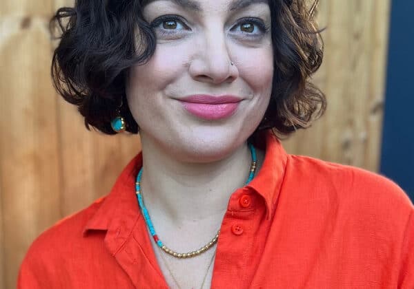 A woman with a dark curly bob smiles past the camera. She has light-toned skin and is wearing a red-orange collared shirt. She has a turquoise necklace and earrings. A wooden fence is in the background.