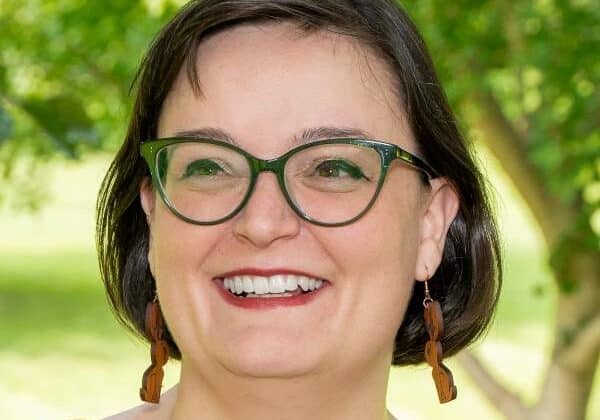 A woman with dark brown bobbed hair and light skin smiles pas the camera. She wears green framed glasses, long earrings, and a dark orange shirt. Green trees and grass are in the background.